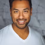 Smiling man with short dark hair and a trimmed beard wearing a white v-neck shirt against a textured gray background.